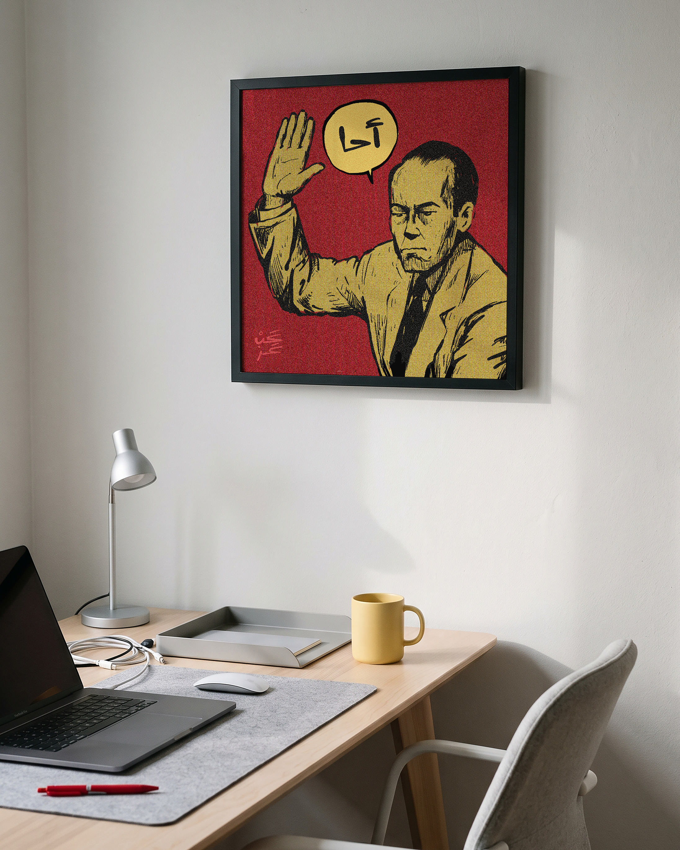 A framed pop-art style poster hanging on a light gray wall above a wooden desk with a laptop, lamp, mug, and chair. The artwork shows a man in a yellow suit against a red background with a yellow speech bubble containing 'Ahha' in Arabic.